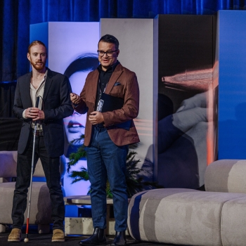 Two presenters stand on stage at The Outlook in front of bold backdrop panels, preparing to speak with microphones in hand.