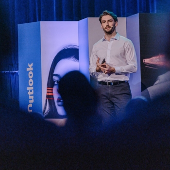 Male speaker framed by audience silhouettes (file-Kpwb53kzw1UsPpWBg2HLVj) Male speaker stands mid-presentation on stage at The Outlook, framed by silhouettes of attendees and panels with striking visual motifs.