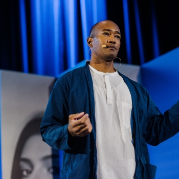 Corey Lee presenting on stage at Design Outlook 2025, wearing a headset mic and gesturing mid-talk against the event’s bold blue backdrop.
