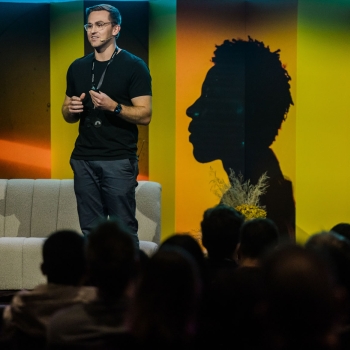 Presenter Matt Perrott speaking on stage at Product Outlook 2025 in Melbourne, standing in front of The Outlook’s signature yellow panels and a silhouette backdrop, addressing an engaged seated audience.