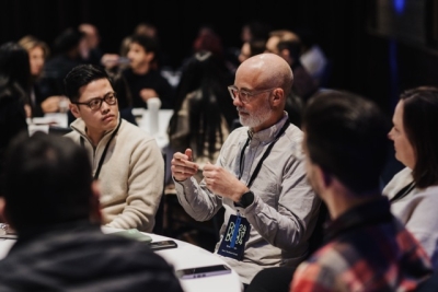 DO24 attendees engaged in a roundtable discussion, with a bearded man in glasses actively contributing while others listen attentively in a collaborative setting.