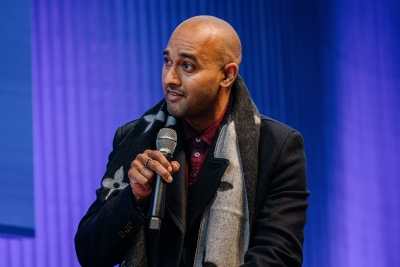 Abi Iyer, a presenter at Design Outlook 2022, speaking into a microphone on stage while wearing a patterned scarf and coat against a purple-lit backdrop.