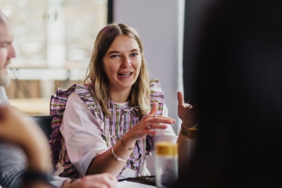 Woman in a ruffled top speaking with animated gestures during a discussion at a roundtable.