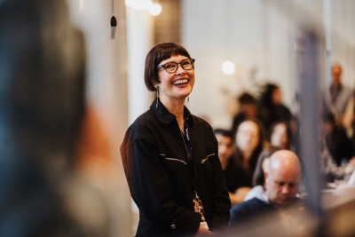Smiling attendee in glasses and a black jumpsuit stands in a warmly lit room at The Outlook, with a softly focused audience in the background.