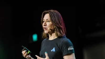 Tarra van Amerongen speaking at PO25, wearing an Atlassian staff shirt, mid-presentation with a mic headset and remote in hand, engaging the audience.