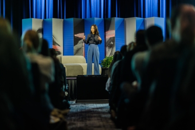 Julia Suh speaking on stage at DO25, framed by striking blue and grey panels, addressing a seated audience in a darkened conference room.