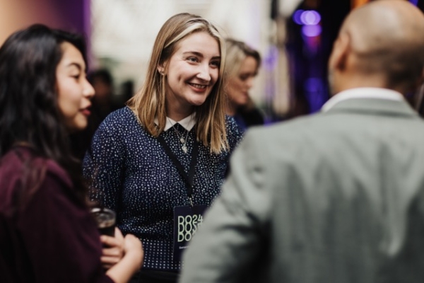 A DO24 attendee, wearing a lanyard, smiles while engaged in a lively conversation with two others in a warmly lit networking space.